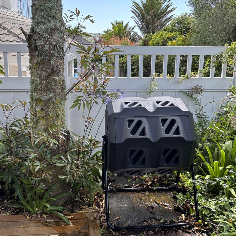 Black compost bin in a garden setting with plants and a house in the background