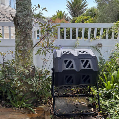 Black compost bin in a garden setting with plants and a house in the background